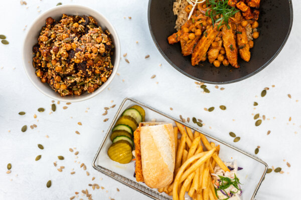Three dishes on a white table: crunchy, grain granola; a fried fish sandwich with pickles and fries; and a curry chicken dish.