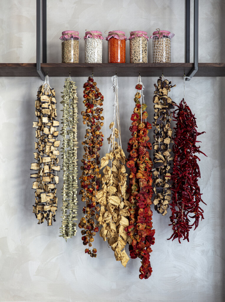 Strings of dried vegetables and peppers hanging beneath jars of grains and legumes, emphasizing sustainable flavor development and traditional preservation methods.