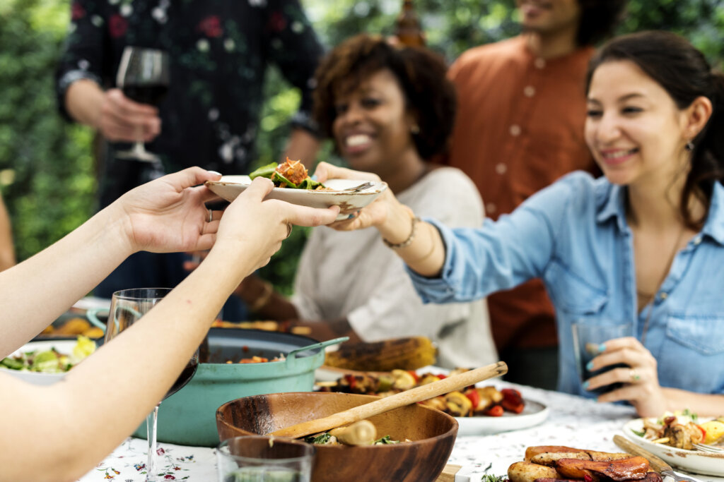 Hands passing a plate of fresh food at an outdoor table with assorted dishes, showcasing authenticity and modern interpretations of traditional flavors.