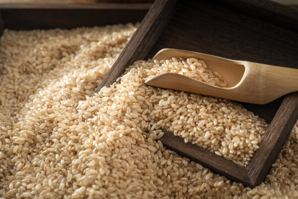 Uncooked brown rice grains piled around a wooden scoop on a dark wooden tray.