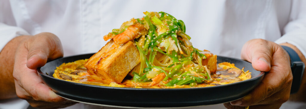 A Griffith Foods chef holding a high protein dish: creamy mushroom omelet with warm silken tofu, dressed with a spicy peanut sesame mala sauce and a green onion and poached shrimp salad.