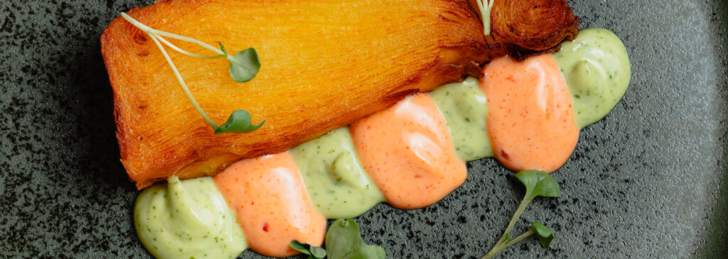 A crispy, fried potato dish served with green and red sauces and microgreens.
