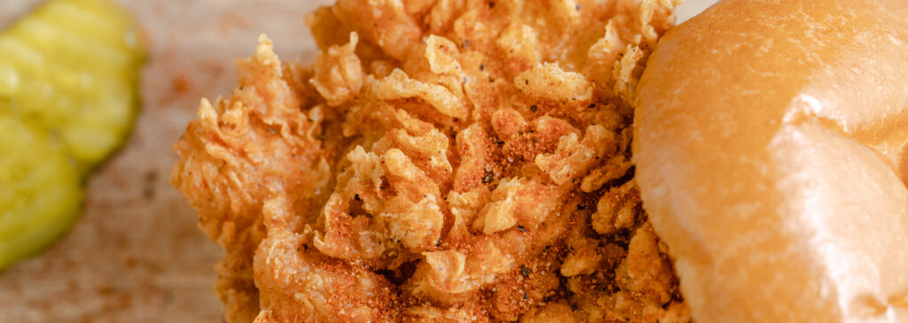 Close-up of a crispy chicken sandwiches’ fried coating, next to a bun and pickles.
